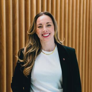 A smiling woman in a white shirt, green pants and black jacket with a Block O lapel pin