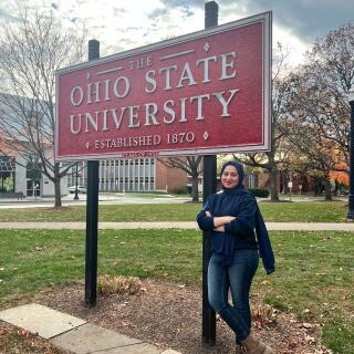 A smiling woman standing with her legs crossed next to a sign reading The Ohio State University